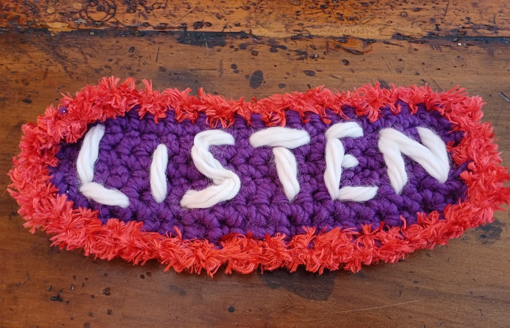 Yarn art with the word 'listen' stitched into it on white thread in all capitals. The background is purple crochet with a red border.