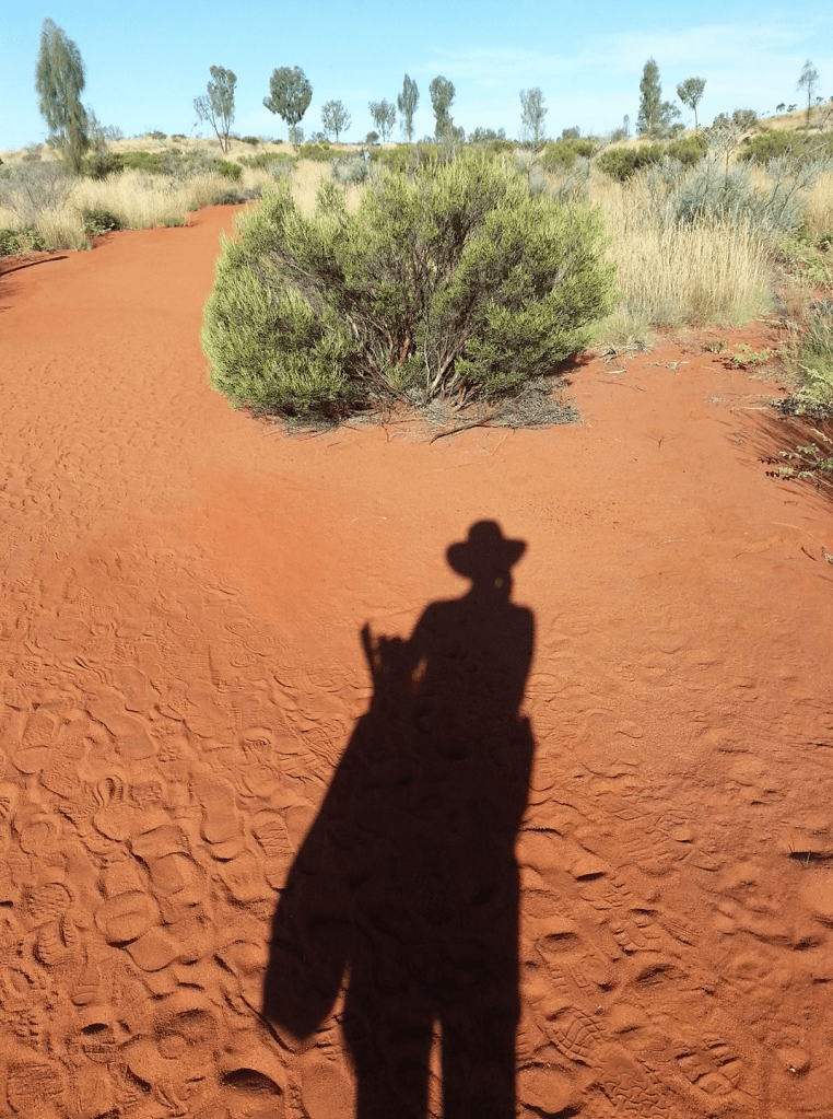 Red sand and low bushes in an arid zone. There is a person's shadow in the foreground, and many different shoeprints in the sand.