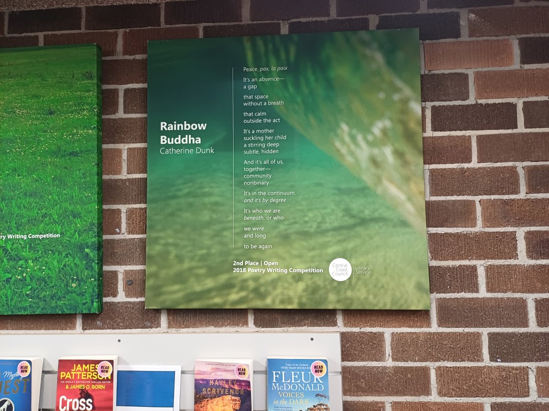 A picture taken in a library that shows the poem printed on a canvas on a brick wall next to a similar canvas and above a shelf of books