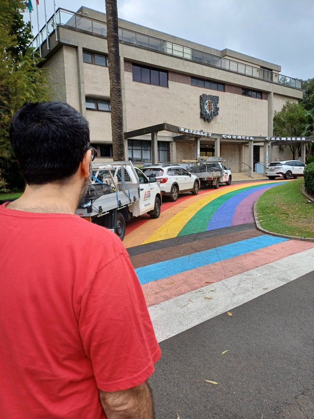 Man in a red tshirt, facing away from the camera, next to a rainbow flag painted on an inner city suburb's road