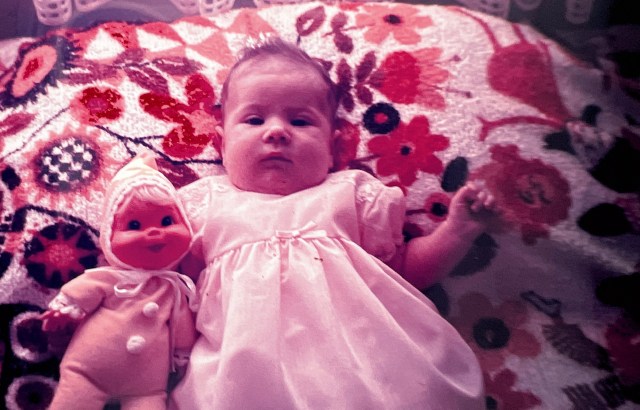 the author as a baby with a doll and lying on a pillow that is from 1970s and garishly patterned