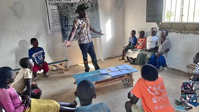 African children in a circle in a bare classroom with an adult speaking to them