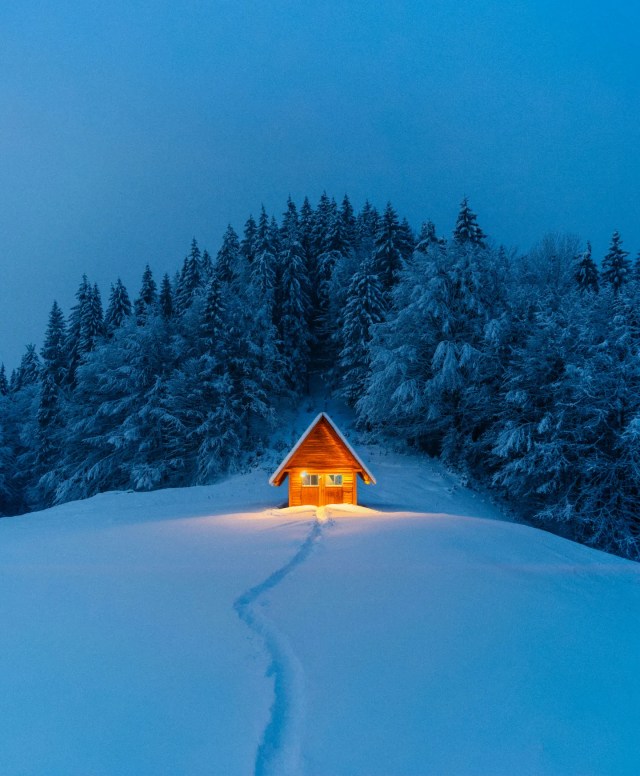 small glowing cabin in the snow in front of pine-covered mountain