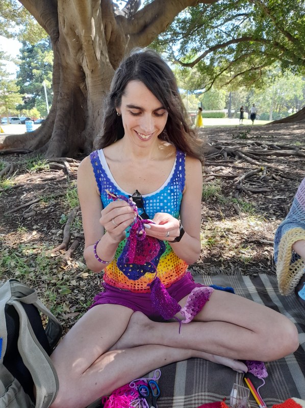 woman sitting on the ground in a park in front of a tree, sewing colourful woollen crochet pieces together