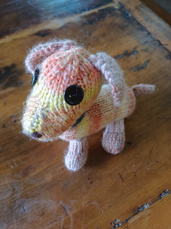 A small knitted toy dog in orange/yellow/beige yarn, standing up on a wooden table
