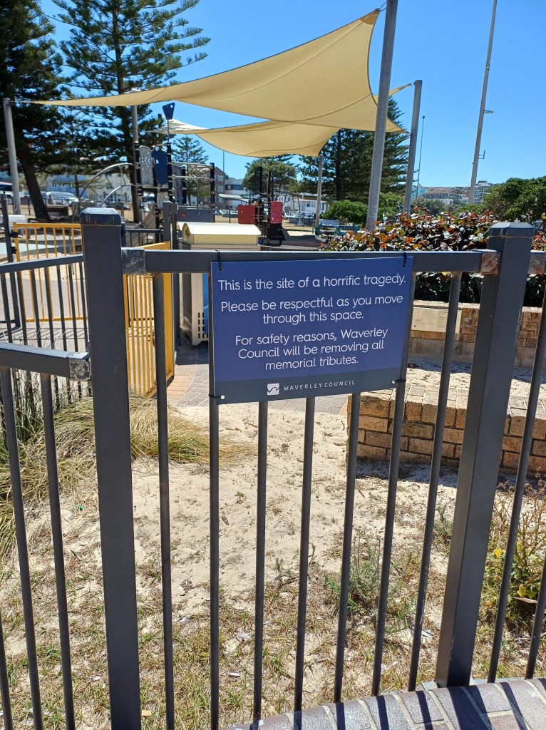 Photo of children's play equipment area at Bondi Beach NSW. A sign on the fence reads: This is the site of a horrific tragedy. Please be respectful as you move through this space. For safety reasons, Waverley Council will be removing all memorial tributes.