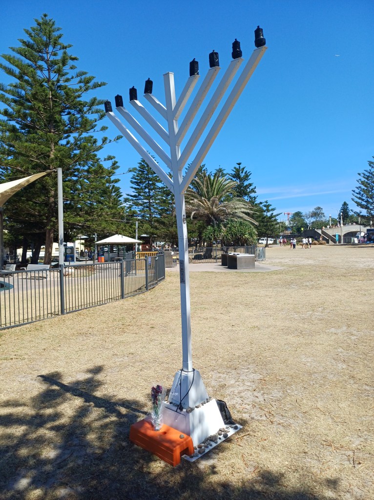 Picture shows a tall metal public menorah in Bondi Beach park New South Wales Australia. There are 8 LED candle type lights, unlit. At the base of the menorah there are a couple of flowers wrapped in plastic and bunched in an orange holder, and there are many small stones placed around the base of the menorah. There is also an unattended pouch or bag partially visible behind the base. An unplugged power cord is wound around the base.