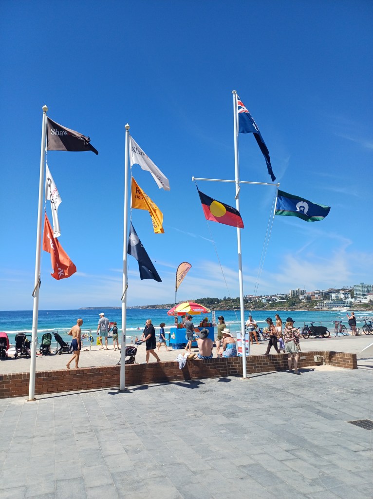 3 flag poles in front of Bondi Beach and the Bondi Surf Lifesaving Club building (not shown). The leftmost two flagpoles feature 3 commercial/advertising flags each. The rightmost flagpole has a cross bar. The main flag is the Australian flag, blue with the British union jack in the corner and the southern cross stars (difficult to see from the photo). The two lower flags on the cross bar are the red, black and yellow Indigenous or Koori flag, and the Torres Strait Islander flag, which is blue, green and white. 2 of the commercial flags, advertising Shaw Partners lawyers, are higher vertically than both of the First Nations Flags, though lower than the so-called Australian flag