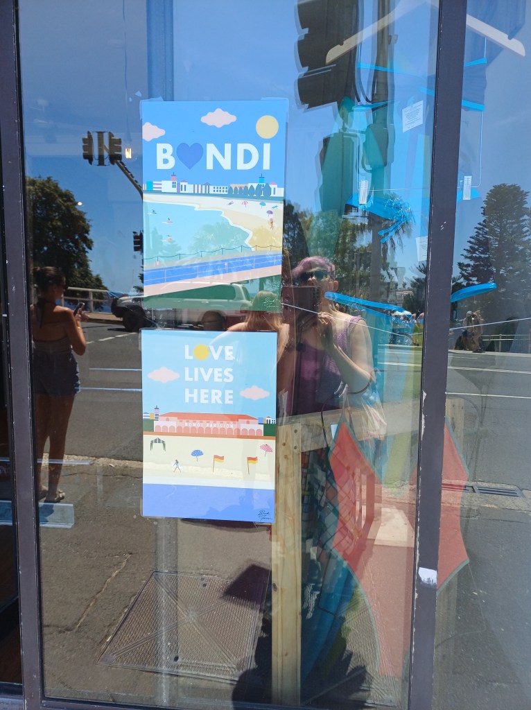 Graphic designed signs in a shop window, reading "BONDI" (the O is a heart) and "LOVE LIVES HERE" (the O is a sun). The photographer is reflected in the window glass. There are other people waiting to cross the road behind her, and a car driving along the road.