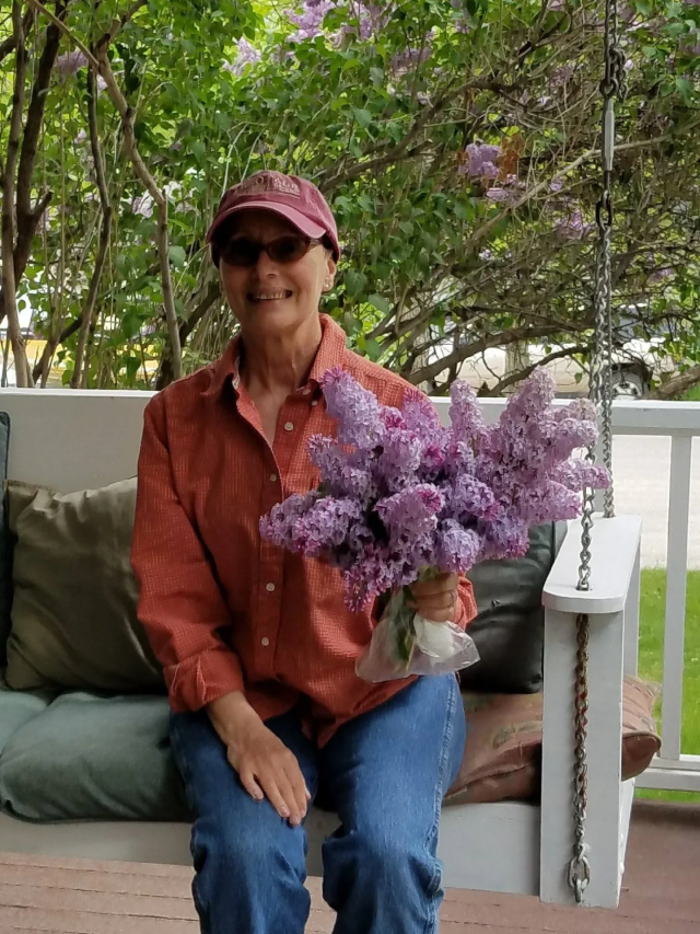 photo of the poet nolcha fox, holding a huge bunch of blooms, sitting on a porch swing. she wears a cap and sunglasses, red long sleeved shirt and jeans