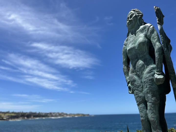 view of mainly blue sky with light cloud and a slim slice of ocean at the bottom. There is a bronze sculpture of a woman at the right hand side, looming large
