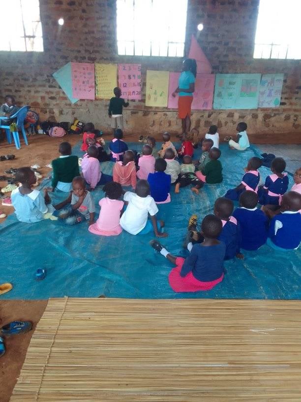 Children learning at a preschool program in kenya. Many children sit on the floor, one boy stands at the front of the room with a teacher. The boy is pointing to a number chart