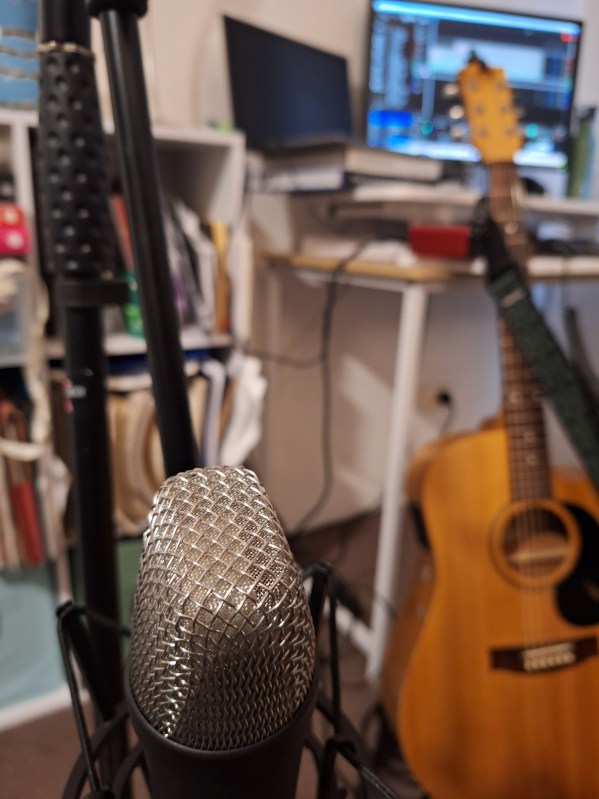 acoustic guitar, microphone and a couple of computers on a small desk in the background, like a home recording studio setup