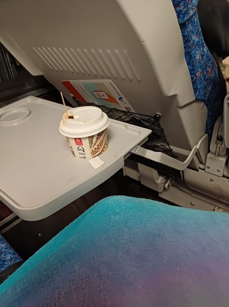 photo taken inside passenger train showing a tray table and a coffee cup and a blanket over knees